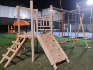 Plazas de juegos de madera modelo Concepción con torre elevada, muro de escalada, escalera y tres columpios de colores.