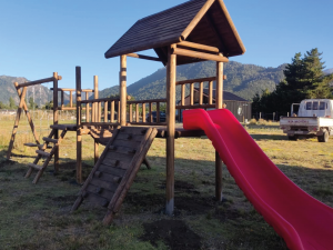Juegos de madera para niños exterior modelo Curacautín con puente de equilibrio, muro de escalada, torre techada y resbalín rojo.