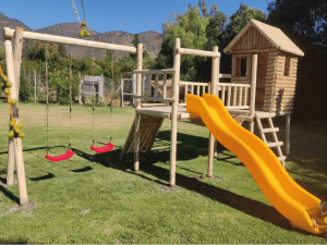 Centros de juegos de madera para exterior modelo Villarrica con cabaña elevada, resbalín amarillo, muro de escalada y columpios rojos.